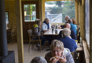 Gasten genieten van eten en drinken aan houten tafels bij Camping De Grienduil, vakantiepark in Utrecht.