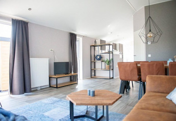 Moderne stue i Waterlodge type 2 med brun sofa, spisebord, reol, tv-bord og store vinduer med gardiner.