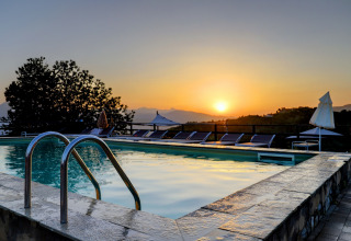 Sunset view over the pool at Camping Pian D'Amora holiday park in Tuscany, Italy, with loungers and mountain scenery.