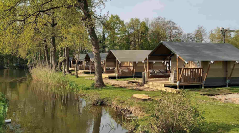Hilera de tiendas glamping junto al agua en Camping Batenstein, Utrecht, Países Bajos, rodeadas de árboles.