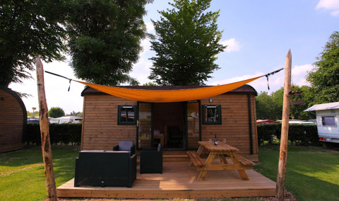 Wooden glamping cabin with deck, outdoor seating, picnic table, and orange sunshade on a sunny day.