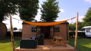 Wooden glamping cabin with deck, outdoor seating, picnic table, and orange sunshade on a sunny day.