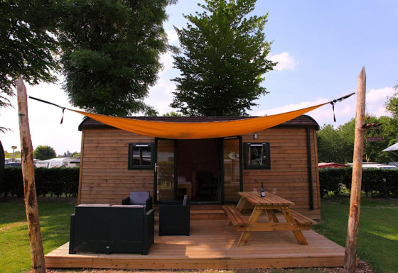 Cabaña glamping de madera con terraza, muebles exteriores y toldo naranja bajo cielo soleado.