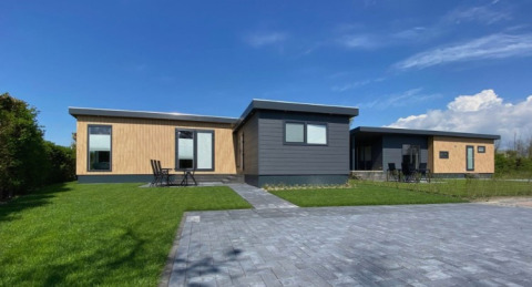 Moderne Lodge De Schelde mit Holz- und dunkler Fassade, Rasen und gepflastertem Weg unter blauem Himmel.