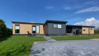Modern lodge De Schelde with wood and dark panels, green lawn, and paved drive under a clear blue sky.