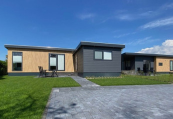 Moderne lodge, De Schelde, med sort og træfacade, grøn græsplæne og stenbelagt indkørsel under blå himmel.