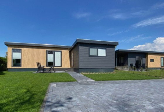 Modern lodge De Schelde with wood and dark panels, green lawn, and paved drive under a clear blue sky.