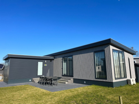 Moderno lodge De Zeester con grandes ventanales, comedor exterior y césped bajo un cielo despejado y azul.