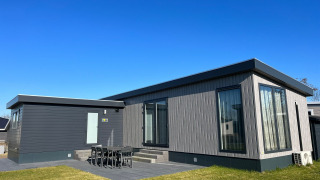 Moderno lodge De Zeester con grandes ventanales, comedor exterior y césped bajo un cielo despejado y azul.
