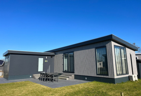 Moderno lodge De Zeester con grandes ventanales, comedor exterior y césped bajo un cielo despejado y azul.