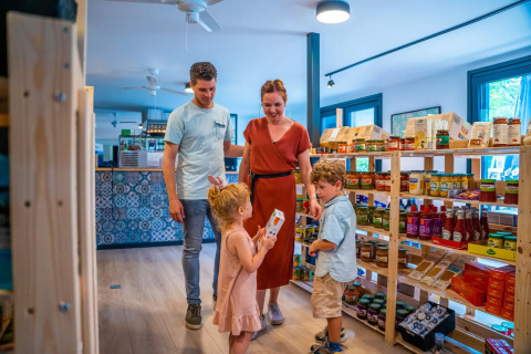 Famiglia che fa la spesa in un negozio di un parco vacanze glamping, tra scaffali di prodotti alimentari.