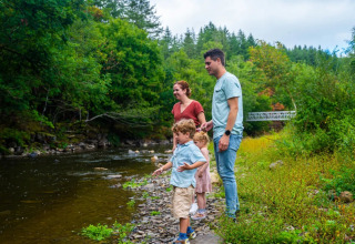 A family enjoys time by a river in a holiday park with glamping, surrounded by lush greenery and trees.