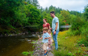 A family enjoys time by a river in a holiday park with glamping, surrounded by lush greenery and trees.