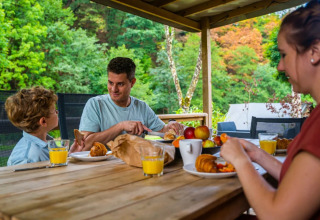 Familie nyder morgenmad udendørs på en terrasse med udsigt til grøn natur i et glamping feriecenter.