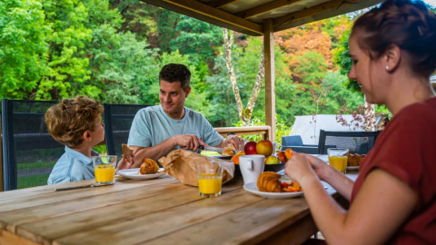 Gezin ontbijt buiten op een houten terras in een glamping vakantiepark met zicht op groen.