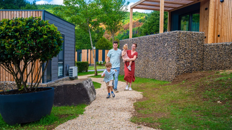 Famille marchant sur un chemin de gravier près de lodges modernes dans un parc de vacances en pleine nature.