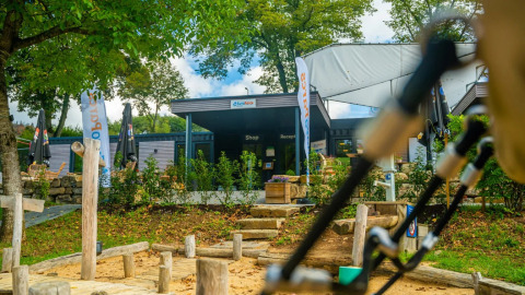 Entrada a un parque vacacional con glamping, tienda y recepción, rodeado de árboles y área de juegos delante.