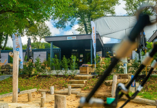 Indgang til feriepark med glamping, butik og reception, omgivet af grønne træer og legeplads i forgrunden.