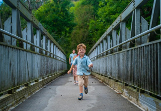 Des enfants courent sur un pont dans un parc de vacances avec hébergements glamping entouré de verdure.