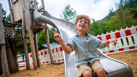 Niño sonriente disfrutando en un tobogán en un parque vacacional con glamping y valla blanca y roja.
