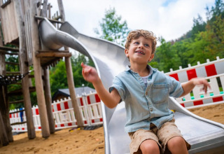 Bambino contento sulla scivolo di un parco vacanze con glamping, recinzione bianca e rossa sullo sfondo.