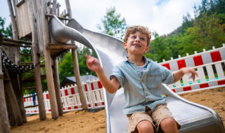 Vrolijk jongetje op een glijbaan in een vakantiepark met glamping, zand en wit-rood hek op de achtergrond.