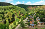 Luftfoto af et feriepark med glamping-hytter, omgivet af grønne skove og en lille flod i dalen.