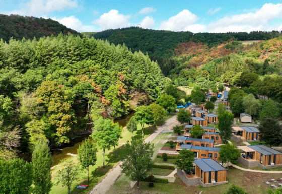 Luftfoto af et feriepark med glamping-hytter, omgivet af grønne skove og en lille flod i dalen.