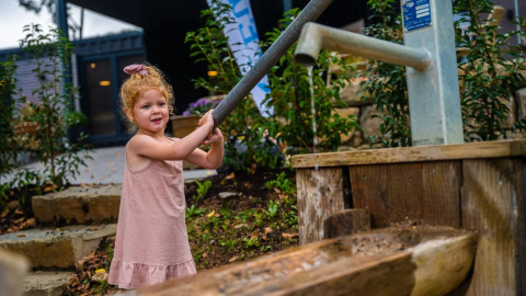 Jonge meid met krullen pompt water bij een buitenpomp op een vakantiepark met glamping faciliteiten.