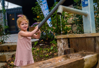 Lille pige med krøllet hår pumper vand fra en udendørs vandpumpe i et feriepark med glamping.