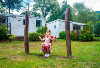 An adult and a child are swinging together in front of modern glamping cabins at a green holiday park.
