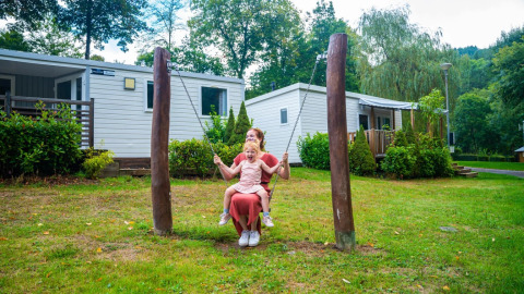An adult and a child are swinging together in front of modern glamping cabins at a green holiday park.