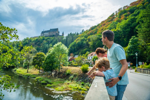 Familie blickt von Brücke auf Fluss und bewaldete Hügel, mit Schloss und Ferienpark im Hintergrund.