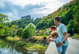 Family enjoys the view from a bridge over a river, with hills, trees, and a castle near a glamping park.