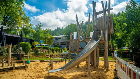 Legeplads med træstruktur og rutsjebane i en feriepark med glamping, omgivet af grønne træer og bygninger.