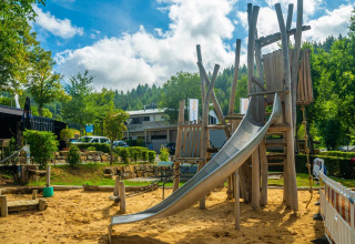 Legeplads med træstruktur og rutsjebane i en feriepark med glamping, omgivet af grønne træer og bygninger.