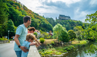 Familia en un puente disfrutando del paisaje de un río, bosque y castillo en un parque vacacional con glamping.