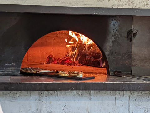 Horno de leña con fuego y pizza dentro, fotografiado en un parque vacacional con glamping.