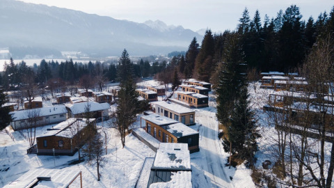 Vista aerea di un parco vacanze glamping innevato con baite in legno tra alberi e montagne sullo sfondo.
