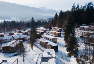 Aerial view of a snowy glamping holiday park featuring wooden cabins amidst trees and mountain scenery.