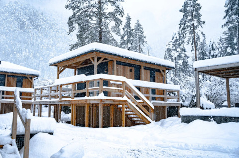 Houten glampingverblijf, bedekt met sneeuw, in een vakantiepark omringd door besneeuwde bomen.