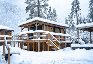 Cabane en bois pour glamping couverte de neige dans un parc de vacances, entourée d’arbres enneigés.