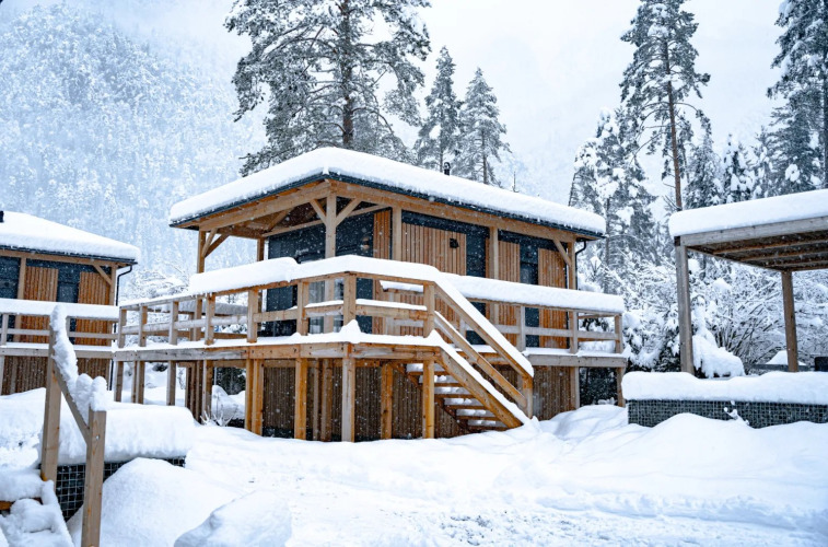 Houten glampinghut bedekt met sneeuw in een vakantiepark, omringd door besneeuwde bomen.