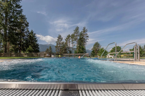 Piscina exterior burbujeante en un parque de vacaciones, rodeada de árboles y montañas al fondo.