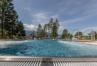 Piscine extérieure bouillonnante dans un parc de vacances, entourée d’arbres et de montagnes au loin.
