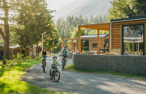Familia pasea en bicicleta por un sendero junto a modernas cabañas de glamping en un parque natural vacacional.
