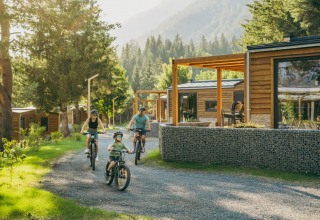 Family cycling on a gravel path past modern wooden glamping cabins at a scenic, forest holiday park.