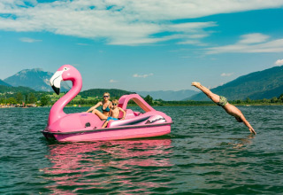 Familie nyder en dag på søen med et lyserødt flamingo-trampolin, mand dykker i vandet, bjerge i baggrunden.