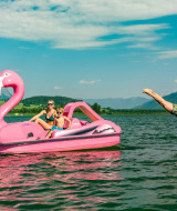 Gezin vaart op roze flamingo-waterfiets op een meer, man duikt het water in, bergen op de achtergrond.