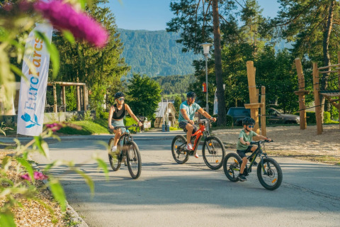 Familie cykler ved et feriecenter med glamping, legeplads og skovklædte bjerge i baggrunden.